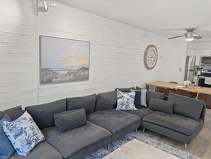 A modern living room with a gray sectional sofa, a wooden dining table, and a kitchen in the background.