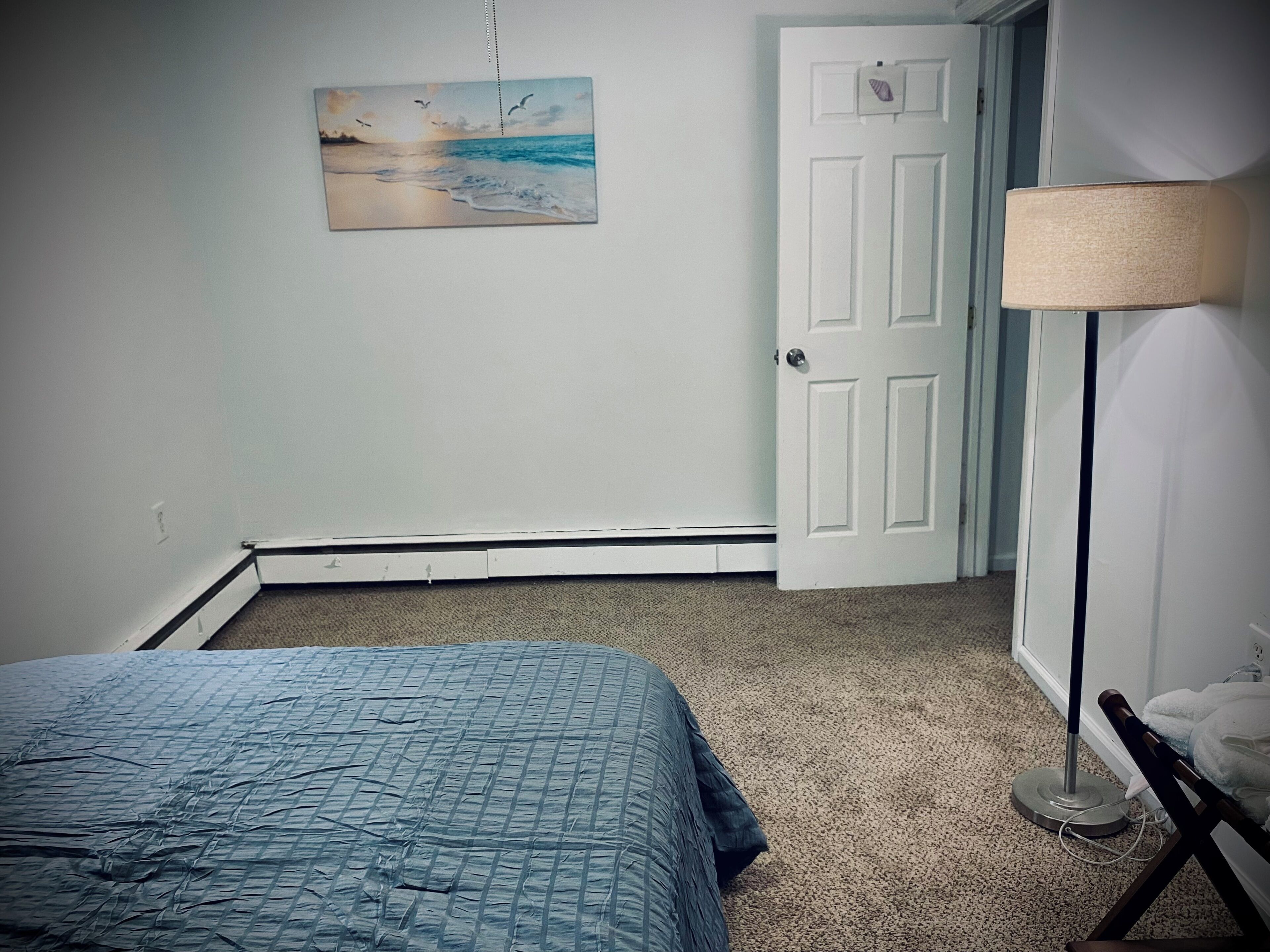 A bedroom with a bed, a painting, a door, and a lamp.