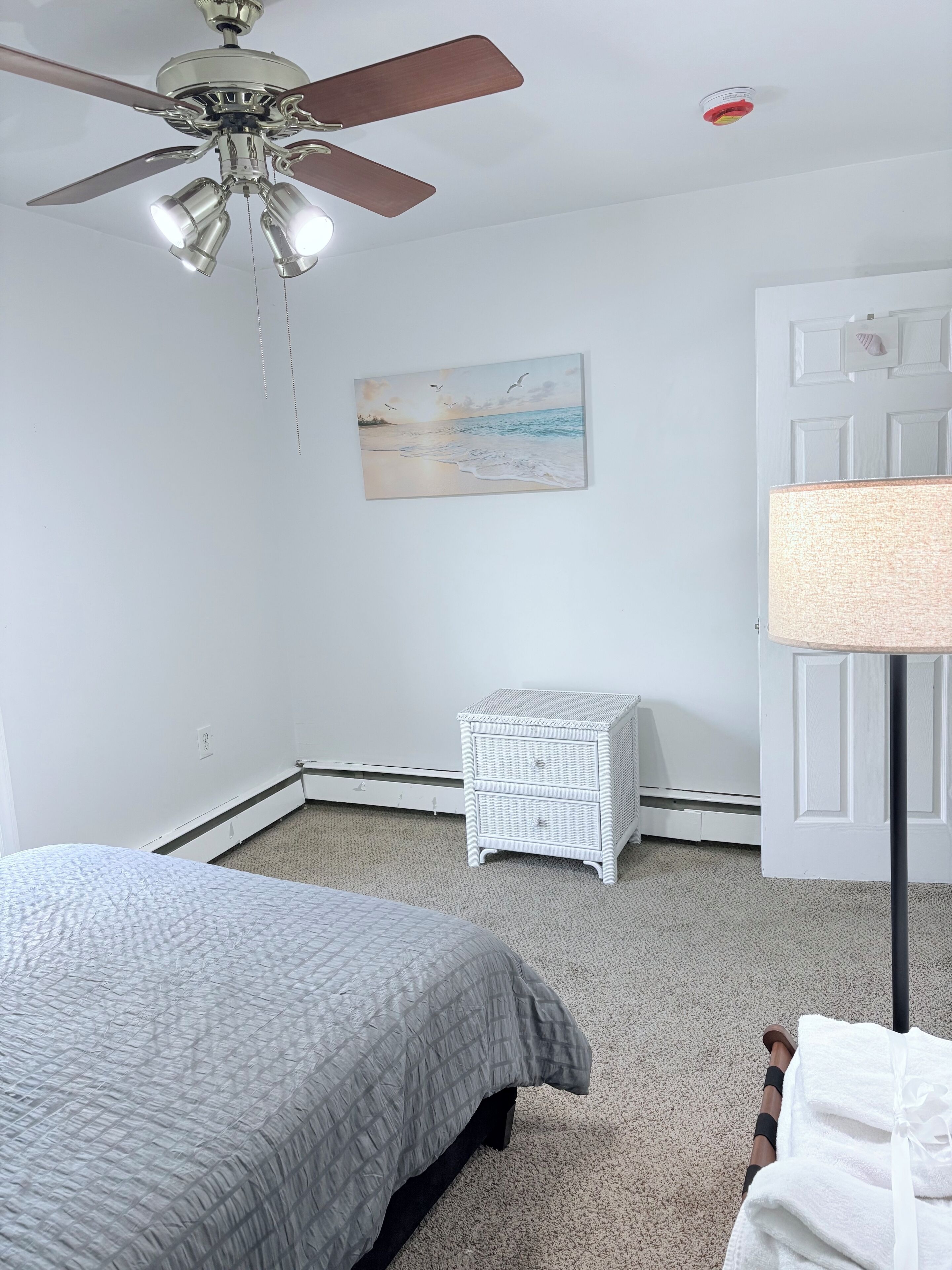 Una camera da letto con un letto, un comodino, una ventola a soffitto, un quadro e una porta.