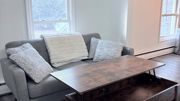 Ein modernes Wohnzimmer mit einem grauen Sofa, gemusterten Kissen, einem Holztisch und großen Fenstern mit Blick auf Bäume.