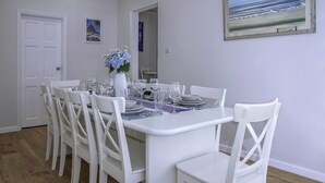 A dining area with a white table set for six, surrounded by white chairs, a vase with flowers, and a painting on the wall.