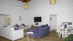 A spacious living area with a white sofa, a purple sofa, a coffee table, and a dining table with chairs. There is a TV mounted on the wall and a plant on the coffee table.
