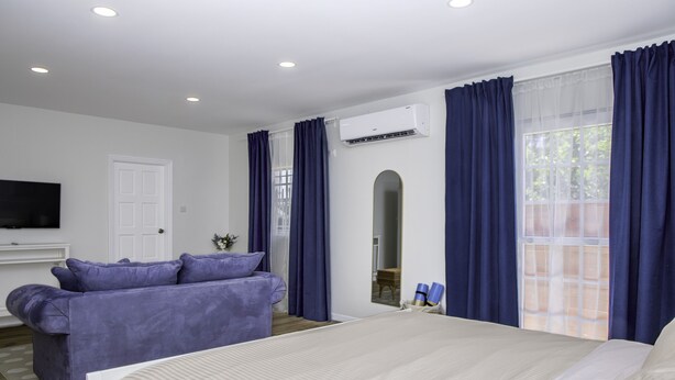 A modern hotel room with a large bed, a sofa, a flat-screen TV, and an air conditioning unit.