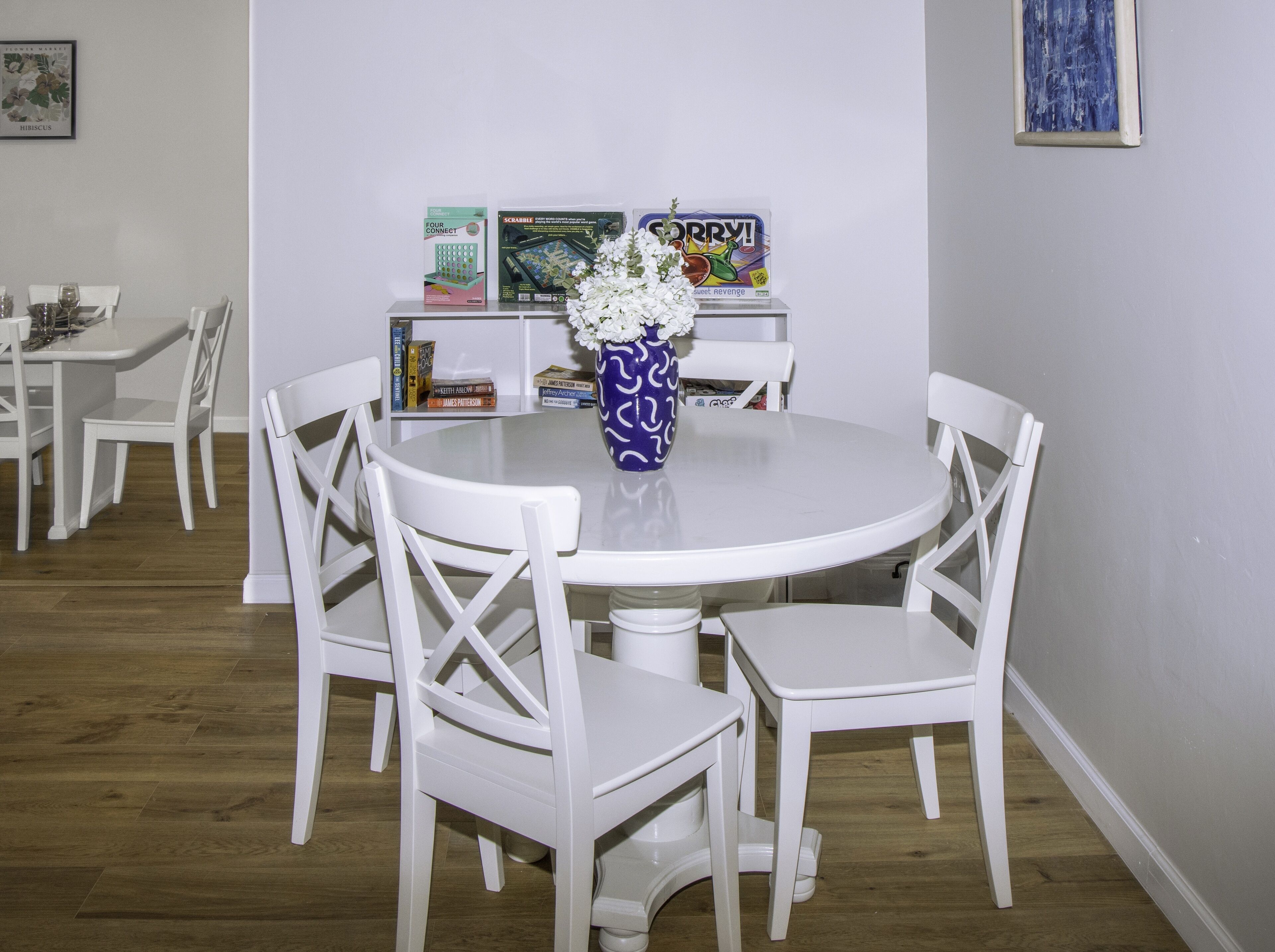 A dining area with a round table, white chairs, and a vase with flowers. There are books and a framed picture on the wall.
