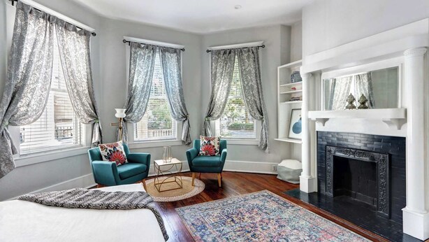 Una habitación con una cama, dos sillones, una chimenea y tres ventanas con cortinas.