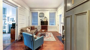 Un salón amplio con sofá, sillón y mesa de centro. Hay dos ventanas con persianas, una chimenea y una puerta con placa de número.