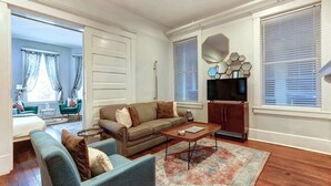 Una sala de estar con un sofá, sillones, una mesa de centro y un televisor. Se observa una alfombra estampada en el suelo.