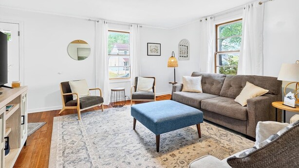 A modern living room with a grey sofa, blue ottoman, and two armchairs. There is a TV on a wooden stand, a round mirror, and a lamp on a side table.