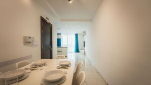 A dining area with a table set for four, a door to the left, and a kitchen in the background. - Gozo Haven With Pool Views in Nadur for Families (Nadur)