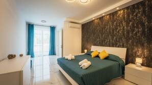 A modern bedroom with a large bed, a nightstand, a desk, and a view of the outdoors. - Gozo Haven With Pool Views in Nadur for Families (Nadur)