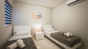 A small, clean room with two beds, a nightstand with a lamp, and an air conditioning unit. - Gozo Haven With Pool Views in Nadur for Families (Nadur)