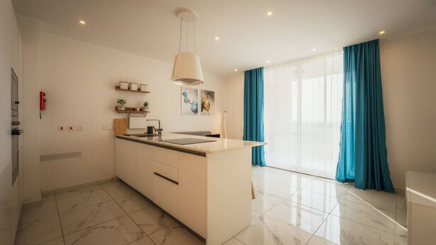 A modern kitchen with a central island, white cabinetry, and a large window with blue curtains. - Gozo Haven With Pool Views in Nadur for Families (Nadur)