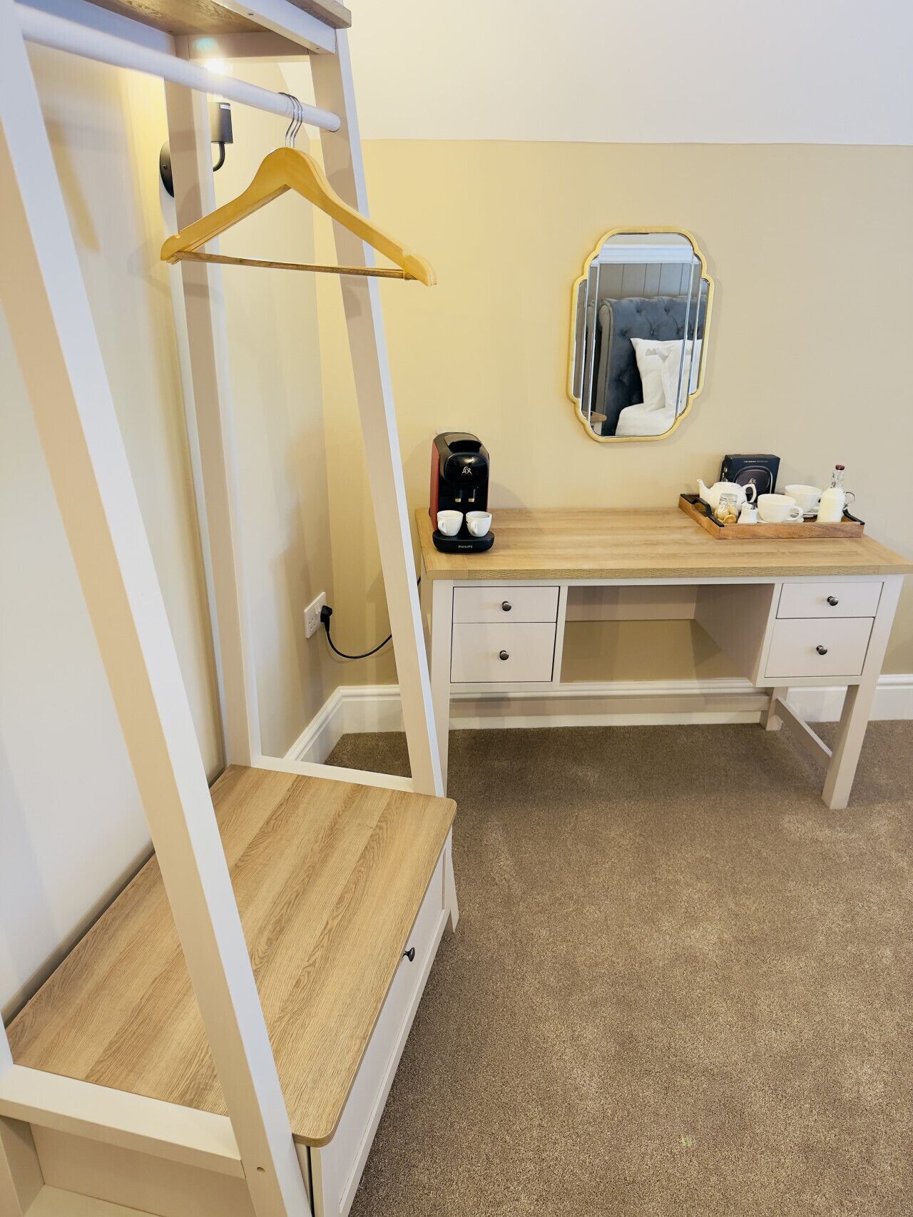 A compact bedroom with a wooden desk, a hanging clothes rack, a mirror, and a coffee maker.