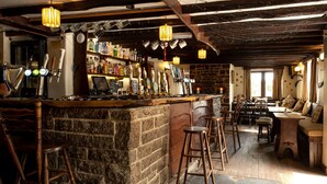 A rustic bar area with a stone counter, wooden stools, and a variety of bottles and glasses. - Rising Sun Inn - Bronn Ewhella (Altarnun)