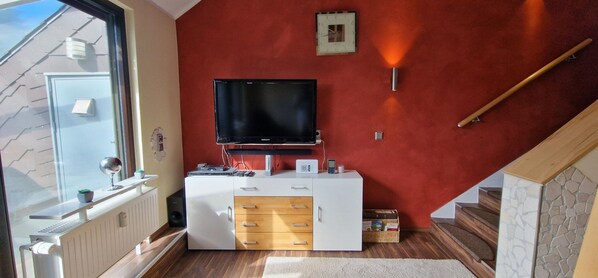Ein kleiner Wohnraum mit einem Fernseher auf einem weißen Schrank, einem Tisch mit einer Pflanze und einem Fenster mit Blick auf ein Gebäude.