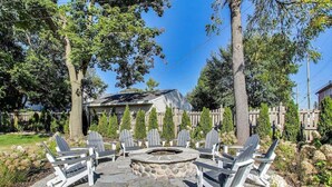 A stone fire pit surrounded by Adirondack chairs in a backyard with a wooden fence and trees.