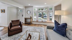 A living room with a brown leather sofa, a gray sofa with a blue pillow, a wooden coffee table, and a large window with a view of the street.