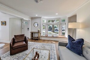 Uma sala de estar com um sofá de couro marrom, um sofá cinza com almofada azul, uma mesa de centro de madeira e uma grande janela com vista para a rua.