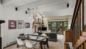 Amplia sala de estar con mesa de comedor de vidrio, sillas blancas, chimenea y ventana grande con vista a una terraza.