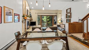 Un comedor espacioso con una mesa de vidrio, sillas blancas y un banco de madera. Se observan cuadros en las paredes, una chimenea y un televisor montado.