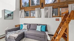 Una habitación con un sofá seccional gris, una manta estampada y una escalera apoyada contra una pared con ventanas amplias que dan a un bosque.