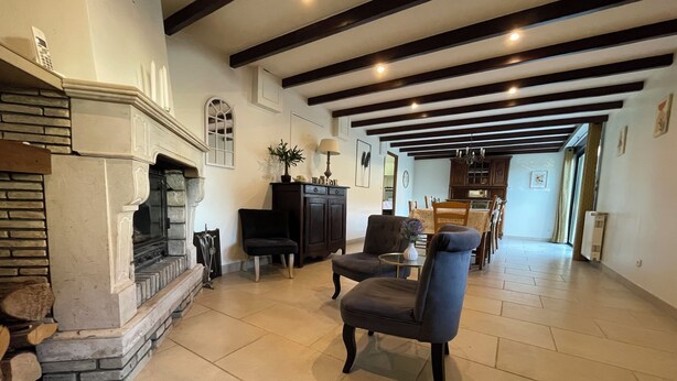 A spacious living room with a fireplace, dark wooden chairs, a dining area with wooden furniture, and a kitchen in the background.