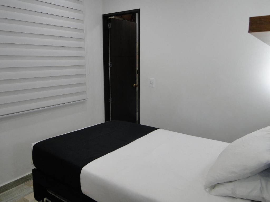 A small, single-bed room with a black and white bedspread, a closed door, and white blinds on the window.