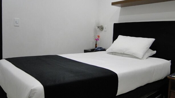 A neatly made bed with a black and white bedspread, a bedside table with a vase of flowers, and a wall-mounted light switch. - Hotel Esmeralda Plaza (Zipaquirá)