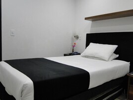 A neatly made bed with a black and white bedspread, a bedside table with a vase of flowers, and a wall-mounted light switch.