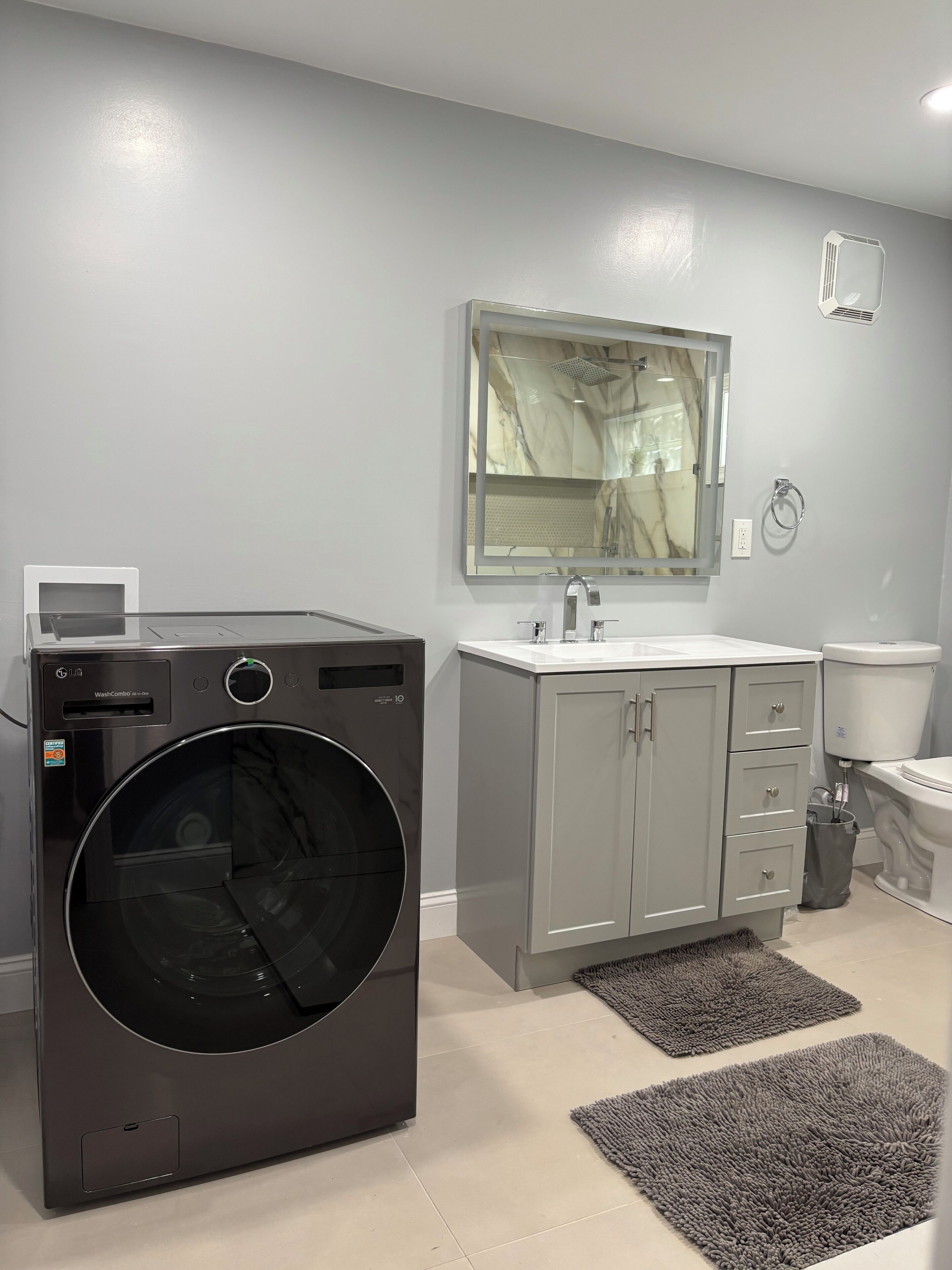 A modern bathroom with a grey sink, a mirror, a toilet, and a black washing machine.