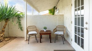 Een overdekt terras met twee rieten stoelen, een bijzettafeltje en een potplant.
