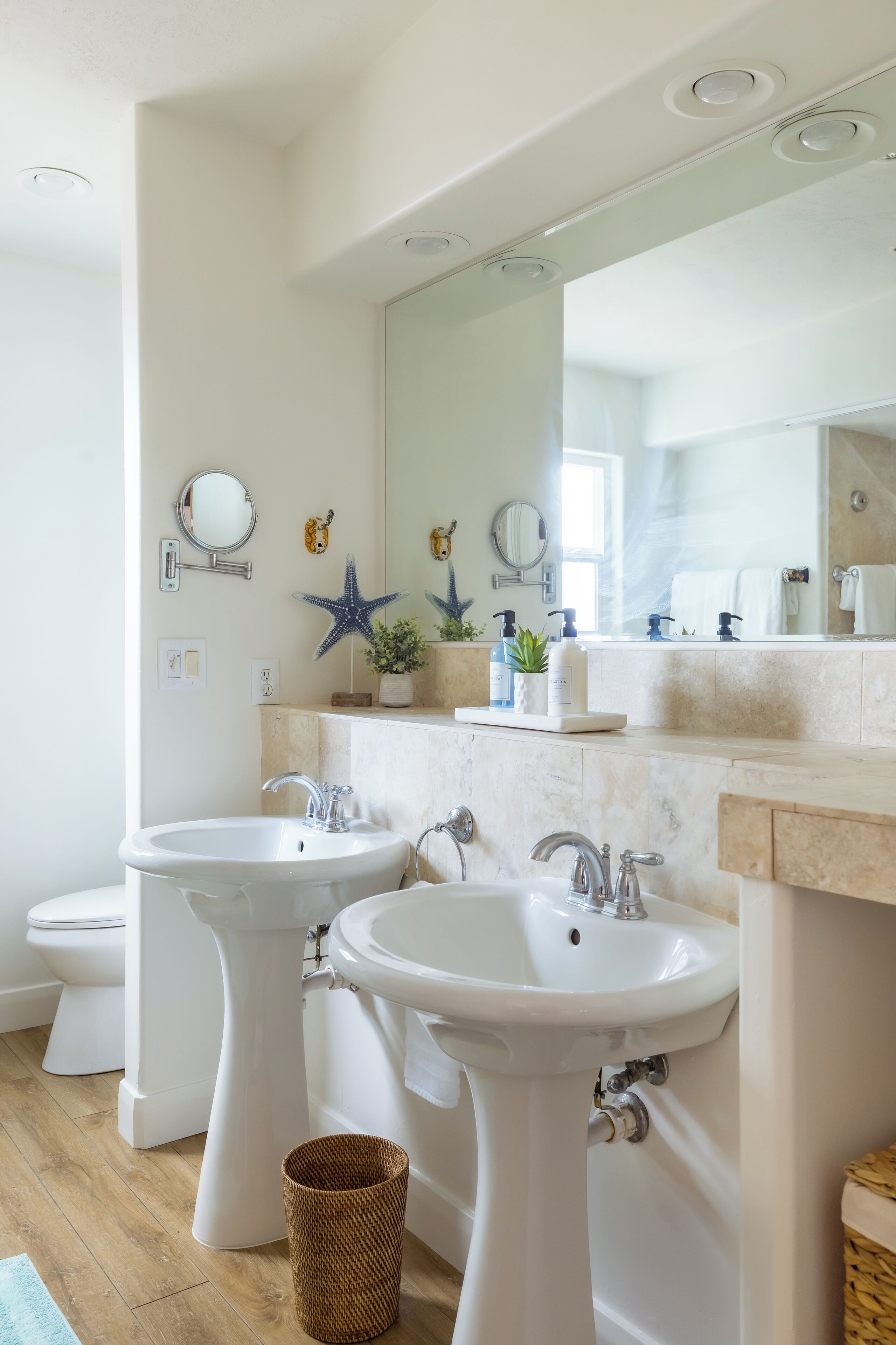 A bathroom with two sinks, a toilet, and a mirror. There are decorative items like starfish and plants on the countertop.