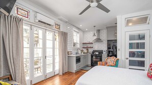 Une chambre avec un grand lit, un plancher en bois, un ventilateur de plafond et une vue sur la cuisine par une porte vitrée.