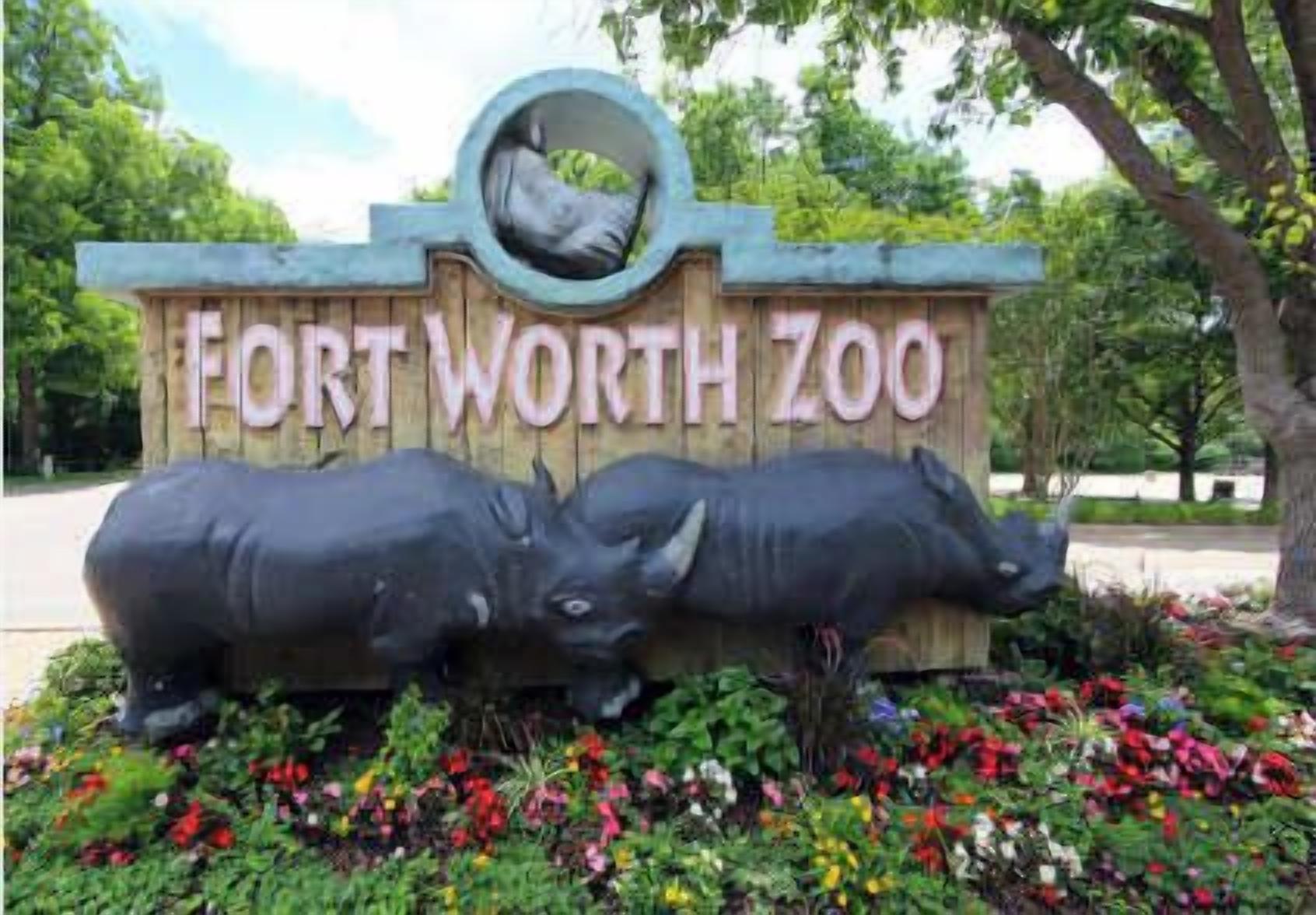 A sign for Fort Worth Zoo with two rhinoceros statues and colorful flowers.