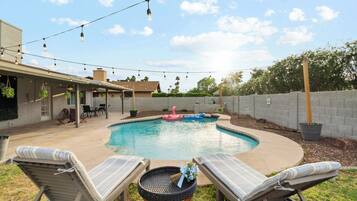 Un patio trasero con piscina, sillas de descanso, luces colgantes y una zona de terraza.