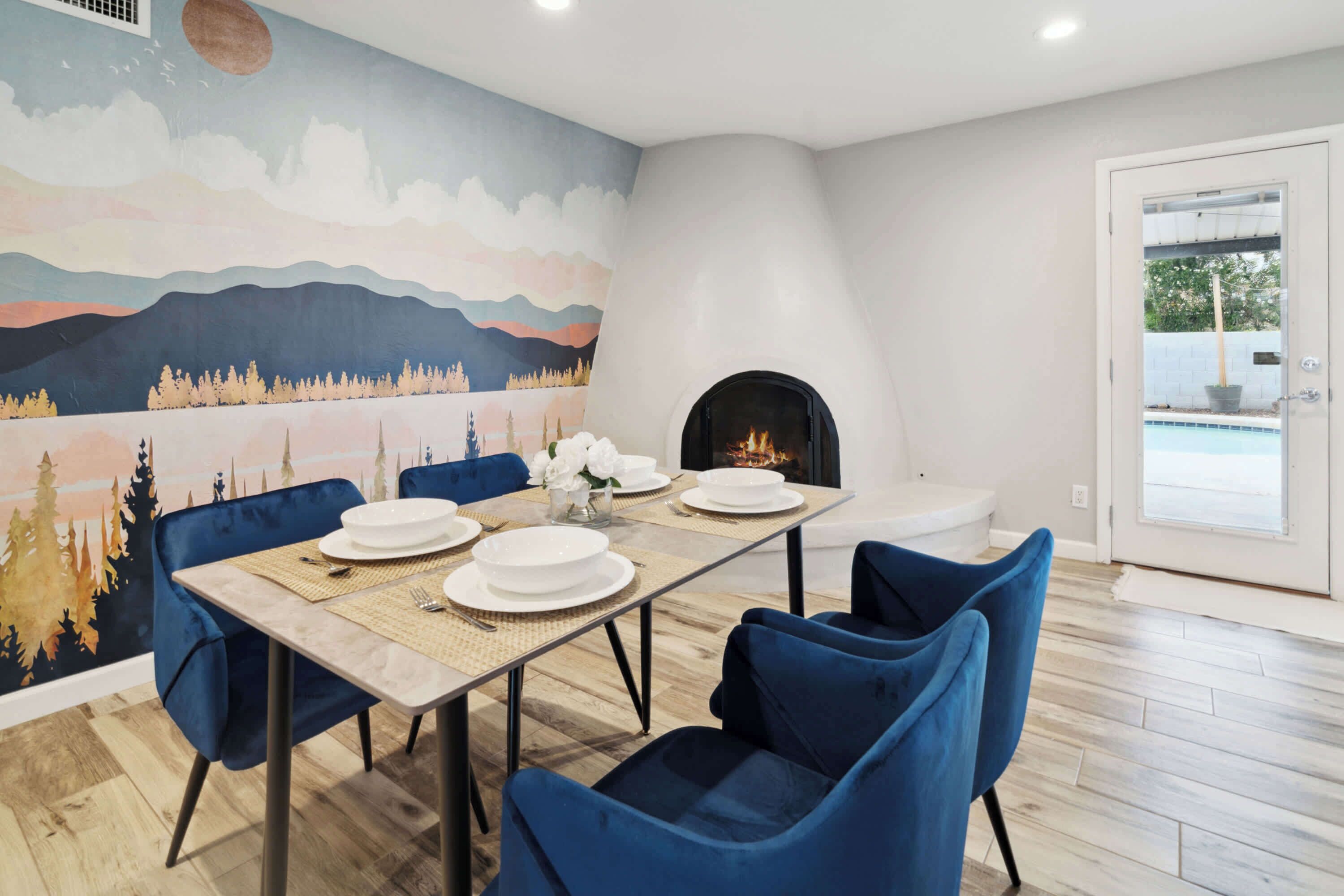 A dining area with a table set for four, blue chairs, a fireplace, and a mural on the wall.