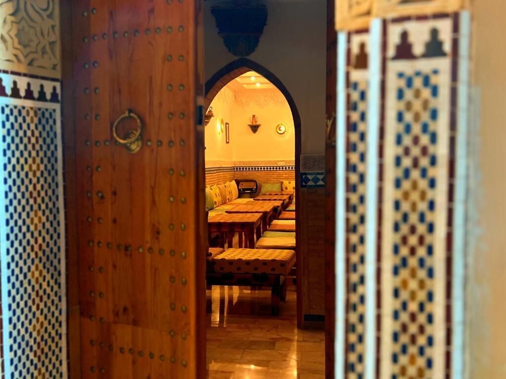 An interior view of a traditional Moroccan riad with a wooden door, a seating area with cushions, and patterned tiles.