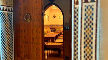 An interior view of a traditional Moroccan riad with a wooden door, a seating area with cushions, and patterned tiles.