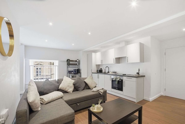 A modern living room with a sofa, a coffee table, and a kitchen area with white cabinets and appliances.