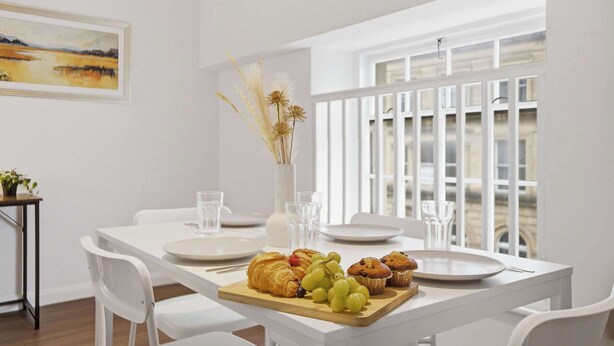 Een eetgedeelte met een witte tafel gedekt voor het ontbijt, met croissants, muffins en druiven.