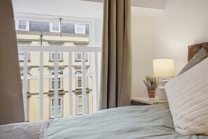 A hotel room with a bed, a nightstand with a lamp, a potted plant, and a view of buildings through a window.