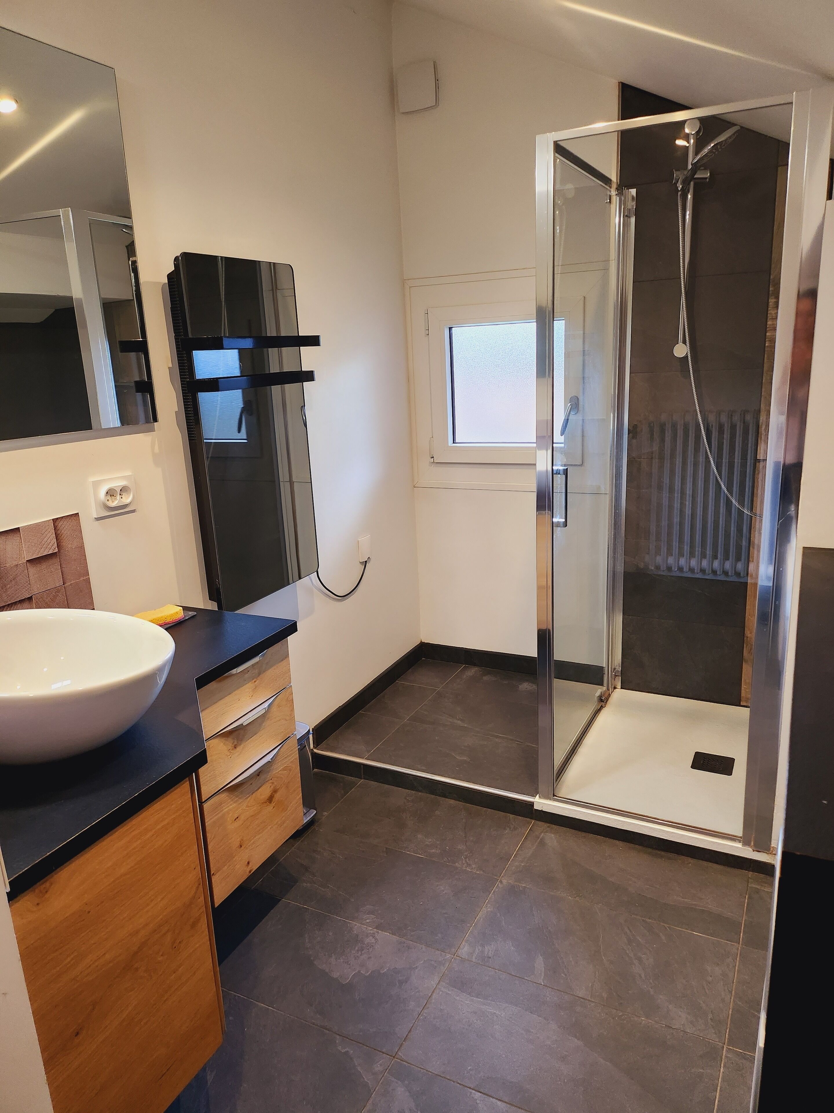 A modern bathroom with a large white sink, a glass shower enclosure, and a window above the shower.
