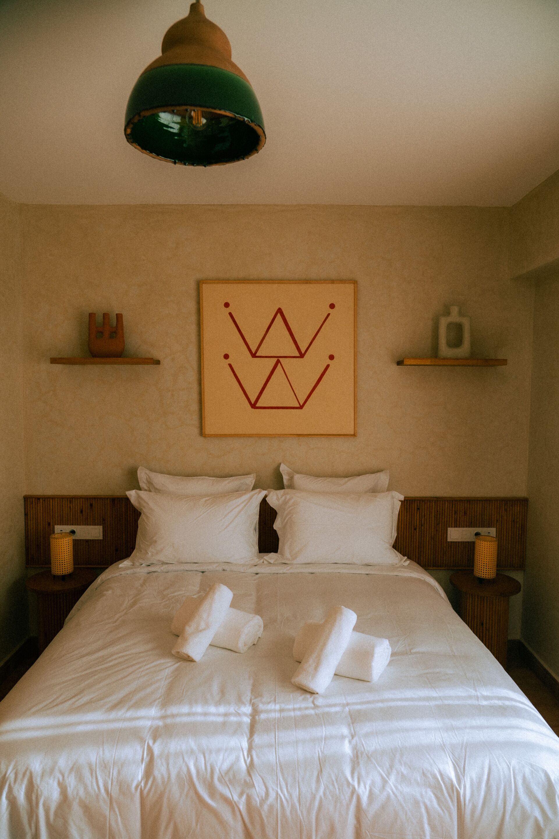 A hotel room with a large bed, two bedside tables, a wall-mounted lamp, and a geometric artwork above the bed.