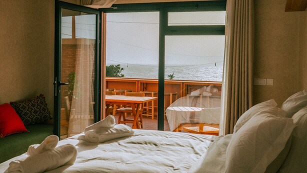 Quarto de hotel com cama, varanda com mesa e cadeiras, e vista para o mar.