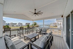 Eine überdachte Terrasse mit Sitzgelegenheiten, einem Deckenventilator und Blick auf ein Gewässer sowie Gebäude.