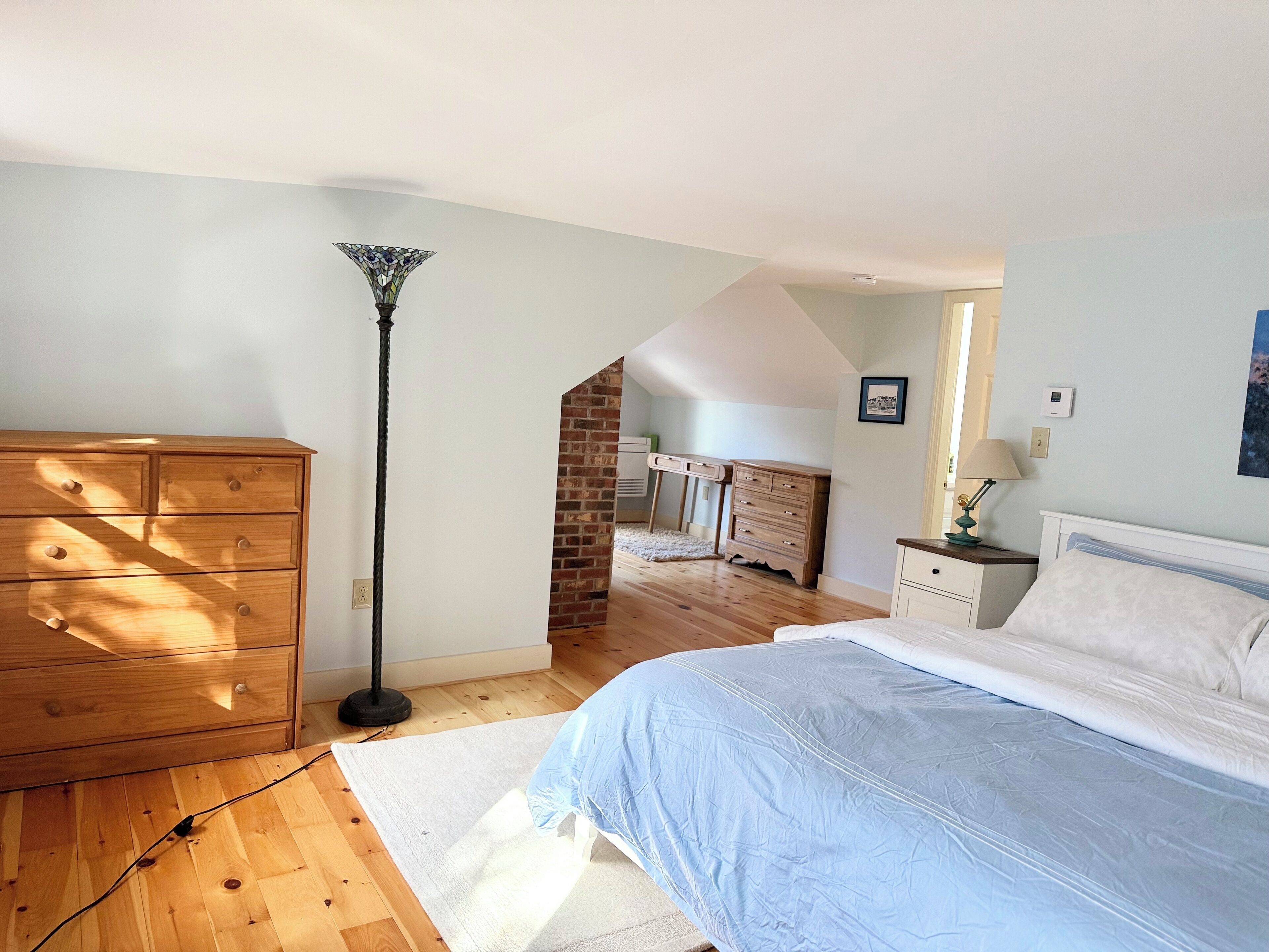 Ein Schlafzimmer mit einem hölzernen Schminktisch, einem Bett mit blauer Bettdecke, einer Stehlampe und einer Ziegelwand.