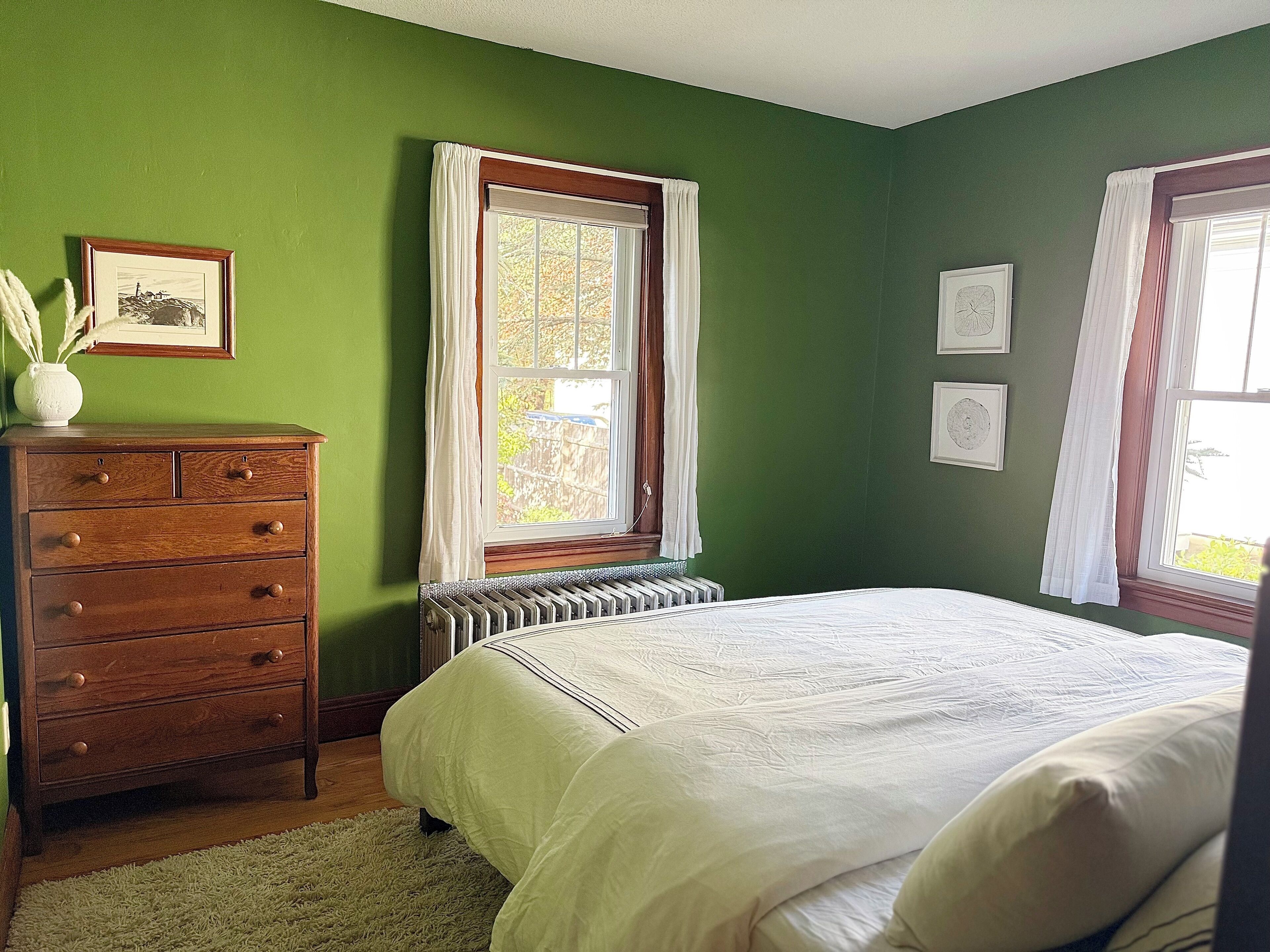 Un dormitorio con paredes verdes, un aparador de madera, un radiador, una cama con ropa blanca y dos ventanas con cortinas.