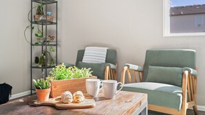 A cozy living room with a wooden table, two green armchairs, a striped pillow, and a plant shelf. - Windshire Retreat | Pool, Garage & Pet-Friendly (Windsor)
