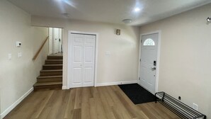 A hallway with wooden flooring, white walls, a black doormat, and a bench. - Lumi Deluxe (Saskatoon)