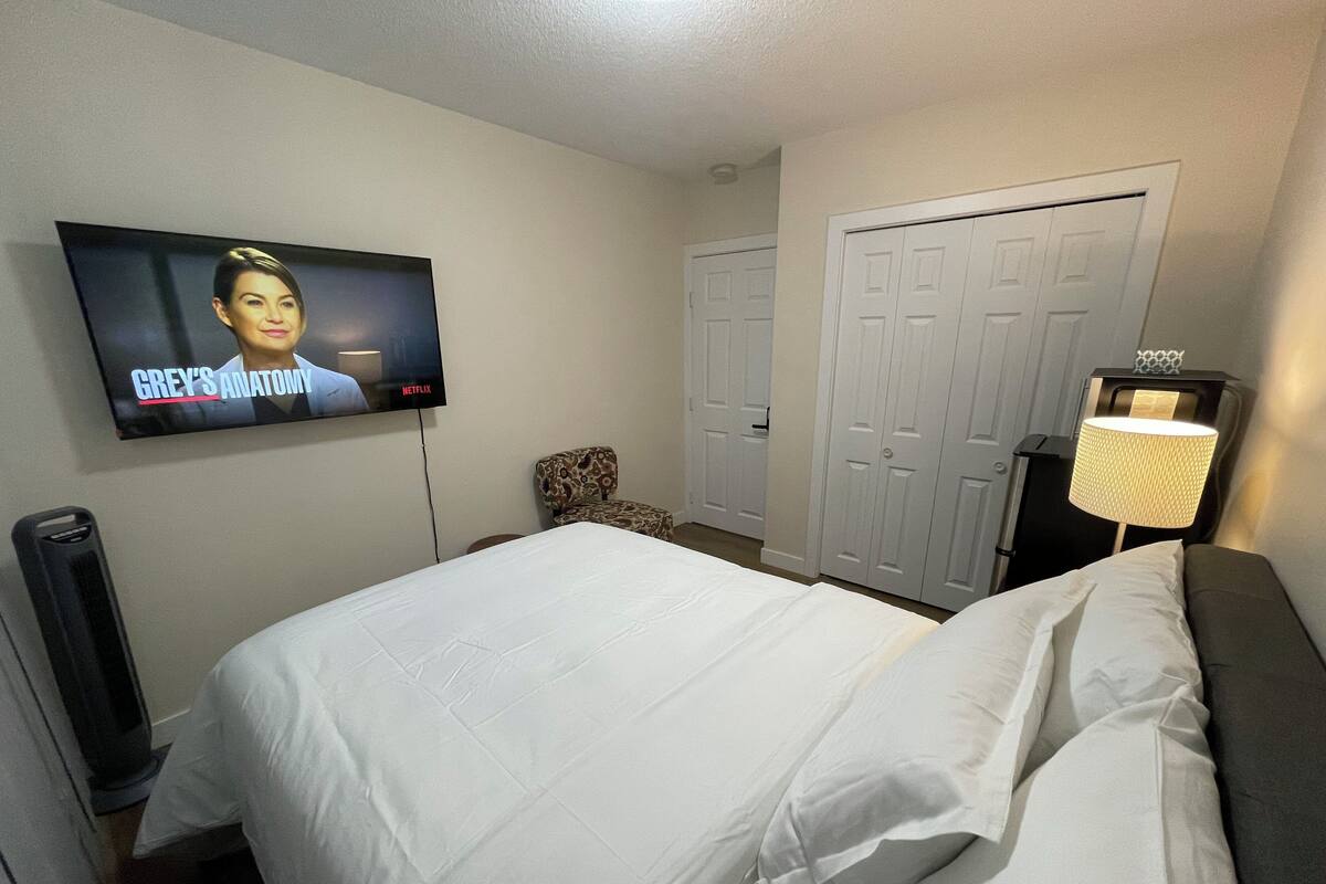 A hotel room with a bed, a TV mounted on the wall displaying 'Grey's Anatomy', a chair, a wardrobe, and a bedside lamp.